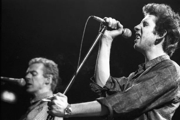 A photo of singer Shane McGowan of The Pogues live on stage.