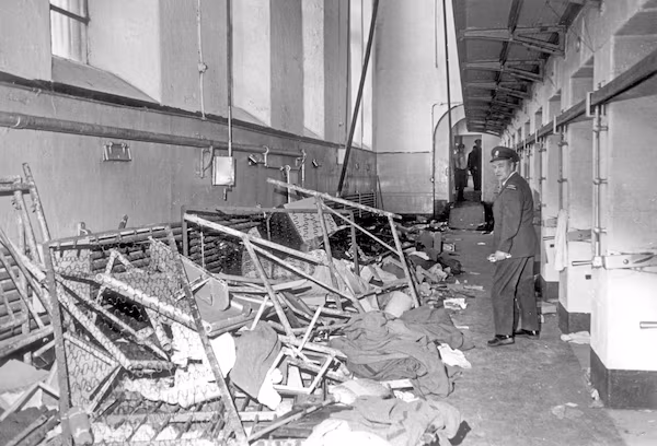 A news photo showing debris from the Kingston Pen. riot of 1971. A guard examines twisted bed frames and ruined mattresses in the hallway of one of the cell blocks that was destroyed.